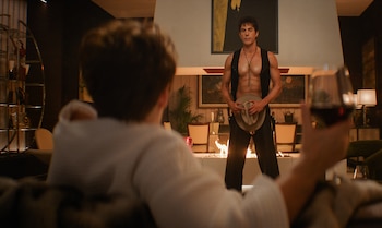 Sacha Baron Cohen, en chaleco y pantalones de cowboy, sostiene un sombrero frente a una chimenea encendida, mientras una persona con copa de vino lo observa