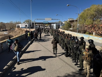 Uniformados con equipamiento táctico alineados en la calzada de un puente junto a civiles y una estructura con un cartel de Bolivia