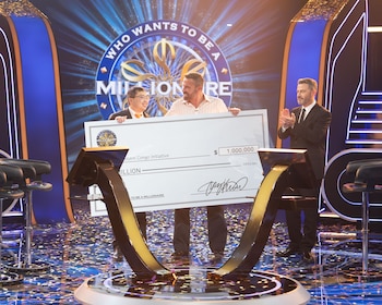 Tres hombres en un escenario sostienen un cheque gigante de un millón de dólares. El fondo muestra el logo de “¿Quién quiere ser millonario?”. Confeti en el suelo