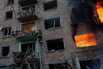 Un bombero asciende una escalera hacia un edificio de ladrillo residencial con ventanas rotas, humo negro y fuego visible en varias unidades
