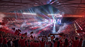 Multitud en las gradas de un estadio con bufandas rojas mientras reflectores y luces multicolores iluminan un escenario.