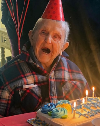 Primer plano de un anciano veterano con un gorro de fiesta rojo, camisa a cuadros, sonriendo y mirando un pastel de cumpleaños con velas encendidas y glaseado azul