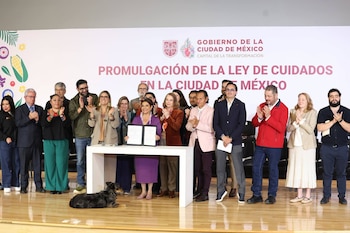Una mujer sostiene un documento frente a un grupo de personas que aplauden. Un letrero al fondo indica 'LEY DE CUIDADOS'. Un perro está en el suelo