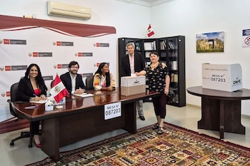 Una mujer deposita su voto en una urna de ONPE, observada por otras cuatro personas en una sala de la Embajada de Perú en Qatar, con una bandera peruana visible