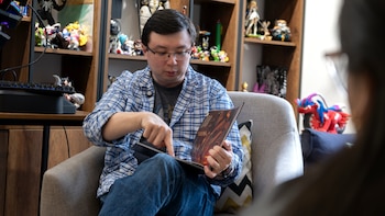 Hombre con gafas y camisa de cuadros, sentado en un sillón, sostiene un libro abierto y lo señala con el dedo. Al fondo, estanterías con figuras