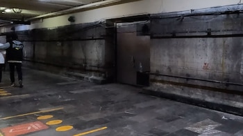 Pasillo subterráneo del Metro CDMX. Muros de concreto, techo bajo con cables y tuberías, suelo de baldosas oscuras con marcas amarillas. Una puerta metálica y dos personas caminando