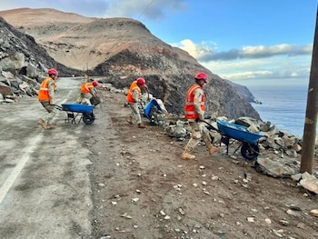 Varias personas con uniformes militares, chalecos reflectantes naranjas y cascos rojos remueven rocas con carretillas en una carretera costera junto a montañas y el mar