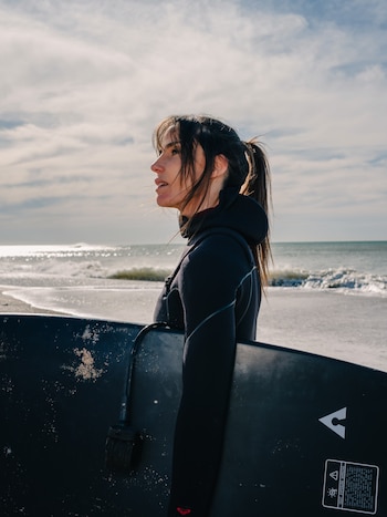 Una mujer de perfil con traje de neopreno negro sostiene una tabla de surf oscura frente al mar bajo un cielo parcialmente nublado