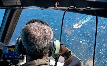 Vista desde la cabina de un avión, mostrando la parte posterior de la cabeza de un piloto con auriculares, mirando un buque en el océano
