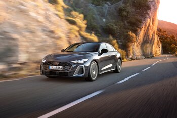 Vehículo Audi gris oscuro en movimiento sobre una carretera asfaltada. A los lados hay montañas rocosas con vegetación y el cielo despejado