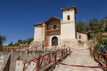 Iglesia de piedra y estuco con torre, balcón de madera, puerta arqueada y techo de tejas. Barandas de madera con cinta roja en primer plano. Cielo azul claro, vegetación