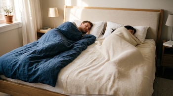 Un hombre y una mujer duermen en una cama cubiertos por mantas de diferente color sobre un colchón con dos almohadas.