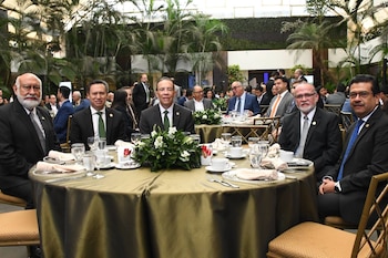 Cinco hombres en trajes oscuros y corbatas están sentados a una mesa redonda con mantel dorado, copas y un arreglo floral