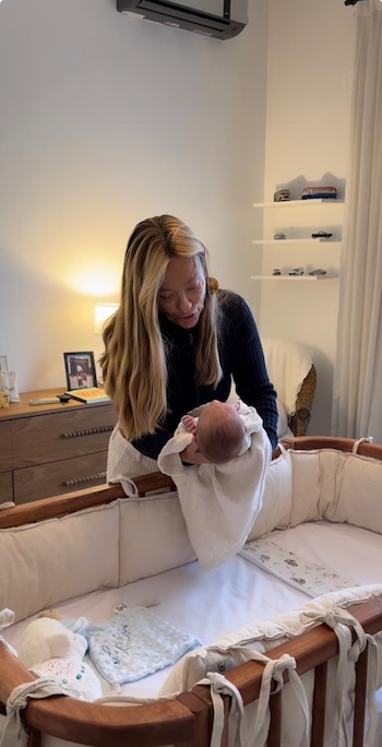 Una mujer con cabello rubio largo sostiene a un bebé envuelto sobre una cuna de madera. En el fondo, un mueble, un aire acondicionado y estantes con autos