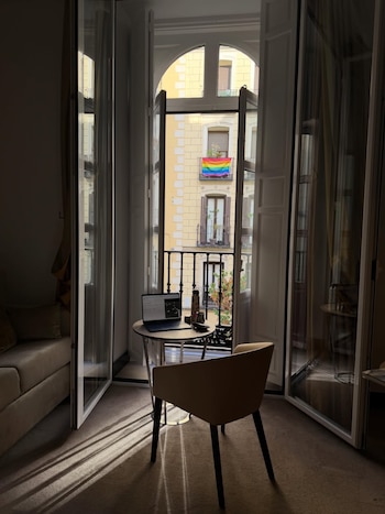 Habitación con puertas abiertas a un balcón. En el balcón de un edificio al fondo, una bandera arcoíris. En la habitación, una mesa con un portátil y un termo
