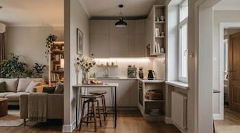 Vista de una cocina pequeña y moderna con gabinetes claros, encimera y barra con dos taburetes, conectada a una sala de estar con sofá y plantas.