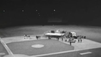 Fotografía en blanco y negro de un campo de béisbol nocturno con un escenario pequeño, personas y vehículos sobre el terreno de juego