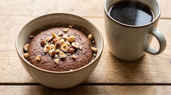 Un tazón con un desayuno vegano oscuro y cremoso, cubierto con trozos de avellanas, se encuentra junto a una taza de mate cocido humeante sobre una superficie de madera.