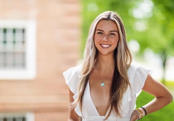 Una mujer joven rubia con vestido blanco sonríe a cámara. Lleva un collar con colgante azul y pulseras en la muñeca. Fondo difuminado de edificio y árboles