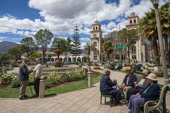 Plaza con fuente, flores, personas mayores sentadas en bancos y de pie. Al fondo, un edificio con torres y andamios, árboles y montañas bajo cielo nublado.