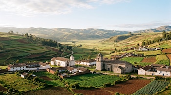 Vista de un poblado con iglesias históricas, casas con tejado y campos de cultivo rodeados por colinas.