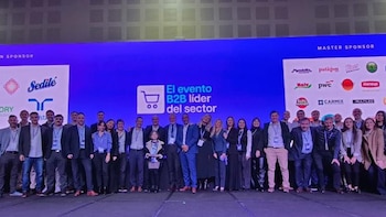 Un numeroso grupo de profesionales posa en un escenario frente a una gran pantalla azul con texto y logos. Una persona al centro sostiene un premio