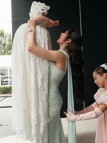Mujer con vestido verde sostiene bebé en traje de encaje blanco con gorro. Niña con vestido rosa y guantes largos observa a la derecha