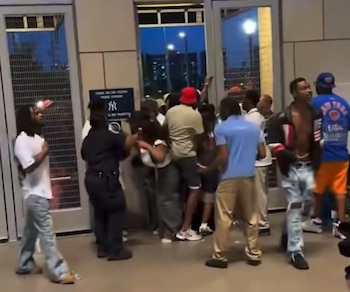 Un grupo de personas se congrega en un acceso del Yankee Stadium de noche. Un agente de seguridad en uniforme oscuro está frente a una mujer