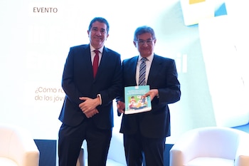 Dos hombres en traje posan de pie frente a una pantalla mientras uno de ellos sostiene un libro con portada ilustrada