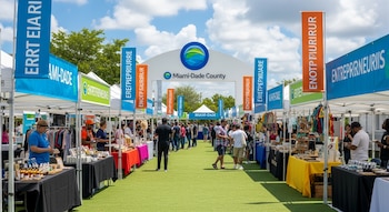 Una feria de negocios al aire libre con múltiples puestos de venta y personas. Un arco de entrada con el logo y nombre de Miami-Dade County es visible.