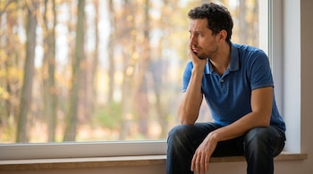 Hombre de cabello oscuro y barba, polo azul, sentado en ventana con mano en barbilla, mirada pensativa. Fondo de árboles otoñales borrosos.