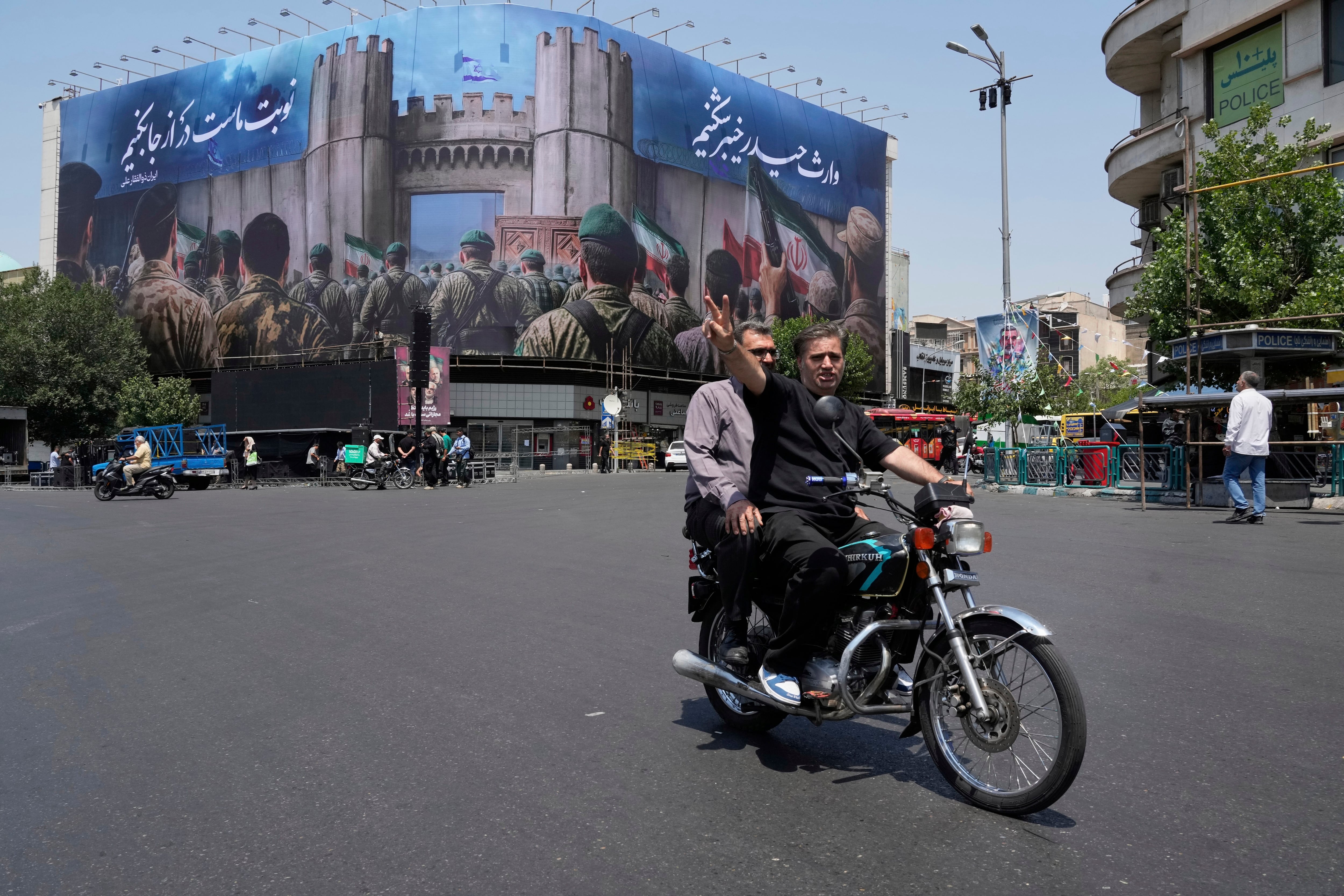 Un hombre hace el signo de la victoria desde su motocicleta tras pasar frente a una manta que muestra a soldados iraníes que se encaminan a atacar territorio israelí, en la Plaza de la Revolución Islámica, el sábado 14 de junio de 2025, en Teherán, Irán (AP Foto/Vahid Salemi)