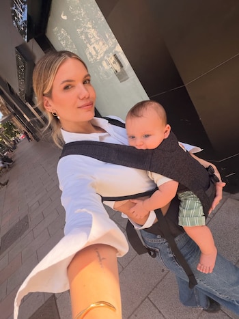 Una mujer rubia sonriente, Eva Bargiela, lleva a un bebé en un portabebés oscuro, Gianluca Simeone, en una calle de la ciudad a la luz del sol