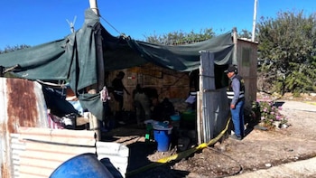 Agentes de la policía y peritos investigan una vivienda precaria con techo de lona verde y paredes de chapa oxidada, rodeada de cinta amarilla de escena del crimen