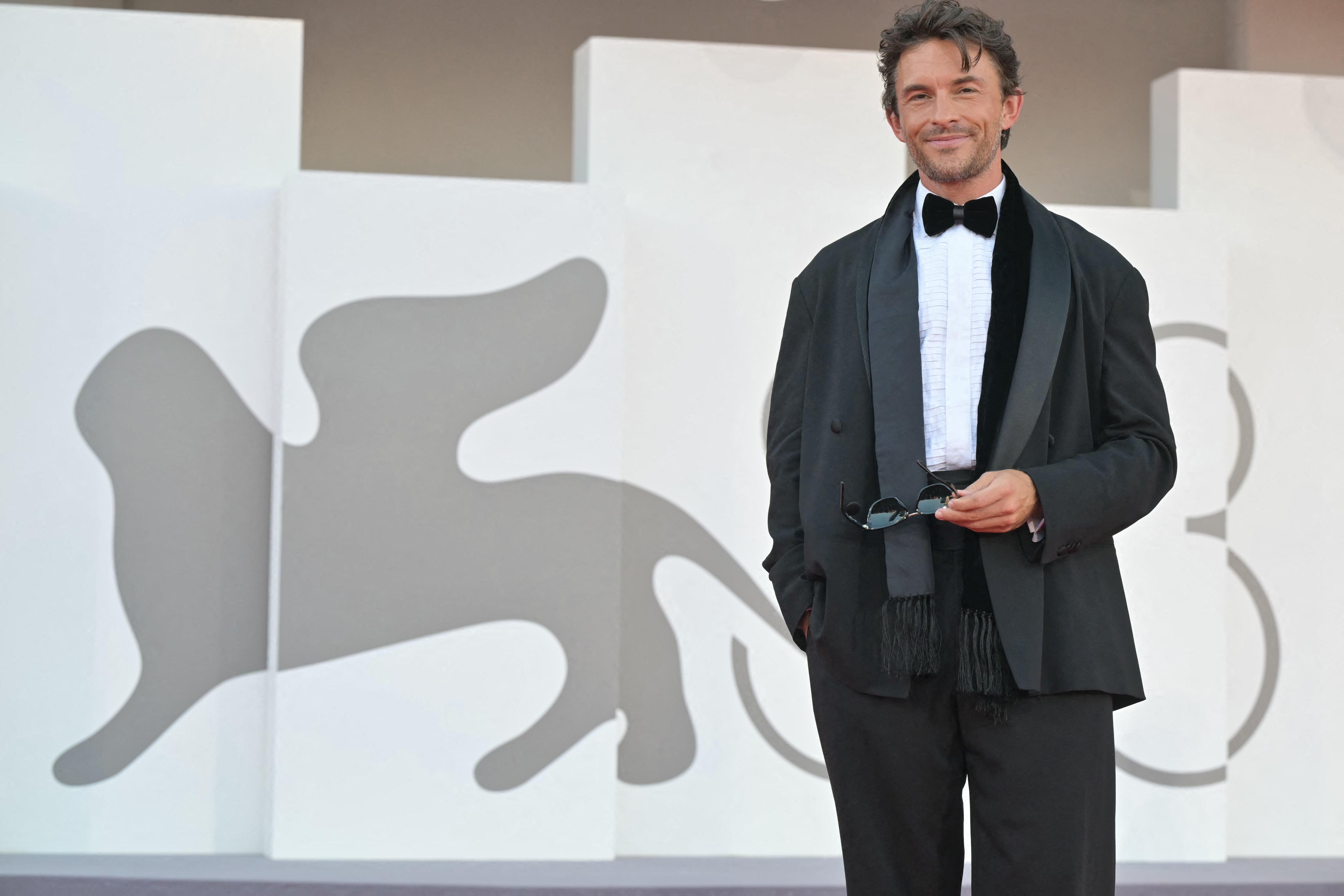 Jonathan Bailey con esmoquin negro, abrigo gris con flecos y bufanda negra (Photo by Stefano RELLANDINI / AFP)