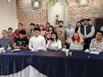 Un grupo de personas en un salón con pared de ladrillo. Una mujer sentada habla por un micrófono mientras sostiene papeles. Hay otras personas de pie y sentadas