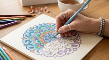 Primer plano de una mano de mujer con anillos y pulsera, coloreando una mandala con un lápiz celeste. La mandala está medio pintada con vibrantes colores.