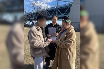 Tres personas, dos hombres y una mujer, participan en una ceremonia al aire libre con un puente y árboles al fondo. El rostro de uno de los hombres está borroso