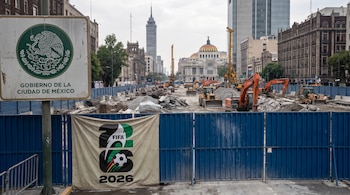 Avenida en obras con vallas azules, excavadoras, cargadoras, escombros, edificios al fondo, cartel del Gobierno de la Ciudad de México y lona del Mundial FIFA 2026.