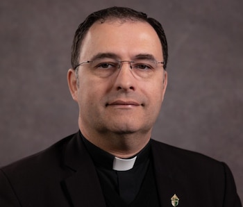 Retrato de primer plano de John Gomez, un hombre de mediana edad con cabello oscuro, lentes y vestimenta clerical negra con cuello blanco
