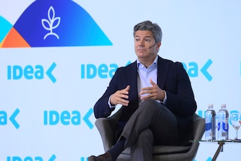 Un hombre con chaqueta oscura y camisa azul habla sentado en un sillón gris frente a un panel blanco con la palabra IDEA