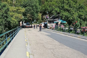 Puente con barandales verdes, una carretera, una acera, árboles, varias personas, un vehículo de emergencia con luces azules, un edificio y flores