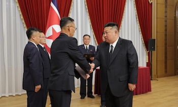 Kim Jong-un estrecha la mano a un hombre de traje que sostiene una carpeta, con la bandera de Corea del Norte y cortinas rojas al fondo