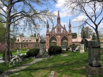 Puertas góticas y edificios de Green-Wood Cemetery con lápidas en un paisaje verde. Árboles en primer plano y la silueta de Nueva York en la distancia