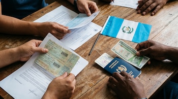 Manos sobre mesa de madera sosteniendo documentos migratorios, pasaportes guatemaltecos, boletos, bandera de Guatemala y billete de quetzal.