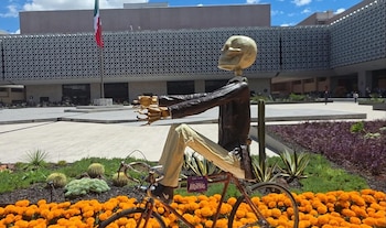 Escultura de un esqueleto en bicicleta sobre flores de cempasúchil con una bandera de México y un edificio de fachada decorada al fondo