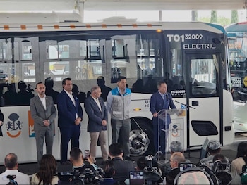 El gobernador Pablo Lemus y funcionarios frente a un autobús eléctrico blanco en un evento. Lemus habla en un podio con micrófono, mientras una audiencia observa