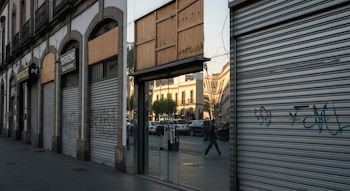 Fachada de edificios antiguos con locales comerciales cerrados por persianas metálicas y paneles de madera. Un cristal refleja la calle, coches y una persona caminando.