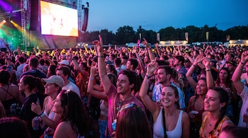 Multitud de personas con las manos levantadas en un concierto nocturno al aire libre. Un escenario con pantalla grande y luces de colores al fondo.