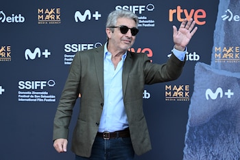 Un hombre con gafas de sol, chaqueta de vestir y camisa azul saluda con la mano izquierda alzada frente a un mural publicitario